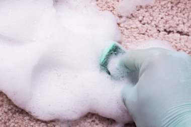 Bol köpüklü deterjanın yanında halı temizliyorum. Yeşil bir süngerle halıyı yıkayan yeşil lastik eldiven verin. Evde bahar temizliği