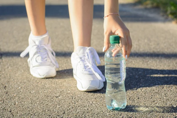 Young sportsmen girl in white sneakers stopped on the road to take a pause and drink some water out of plastic bottle during running in the morning in the countryside