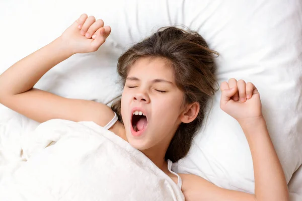 Small girl yawning on comfortable bed in the morning. Little lovely girl wants to sleep in the morning and doesn\'t want to go to school. Concept of yawning and desire to sleep