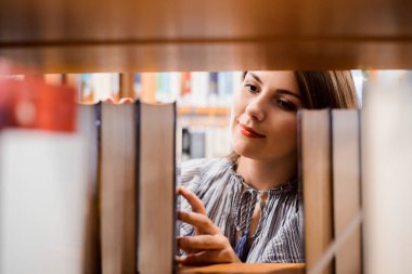 Kütüphanede, farklı kitapların olduğu kitaplıkların yanında duran ve kitaplara göz atmak için kitap alan bir üniversite öğrencisi.