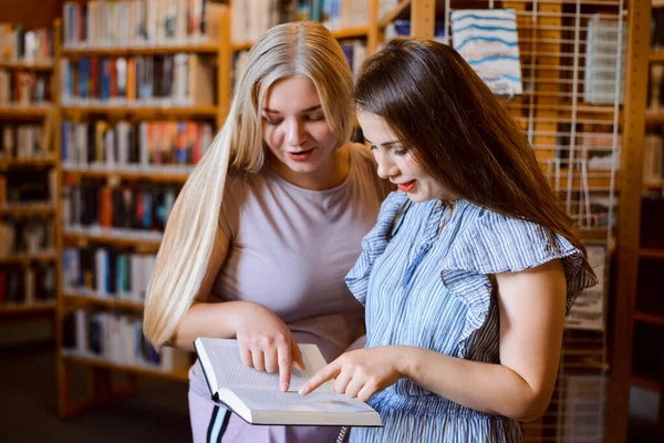 Two girls uni students in library discussing plot of a novel, standing between bookshelves airing views and impressions about the book