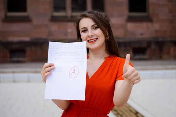 Happy beautiful girl student in red dress showing paper with good test result to the camera boasting about receiving excellent A grade and having high academic performance