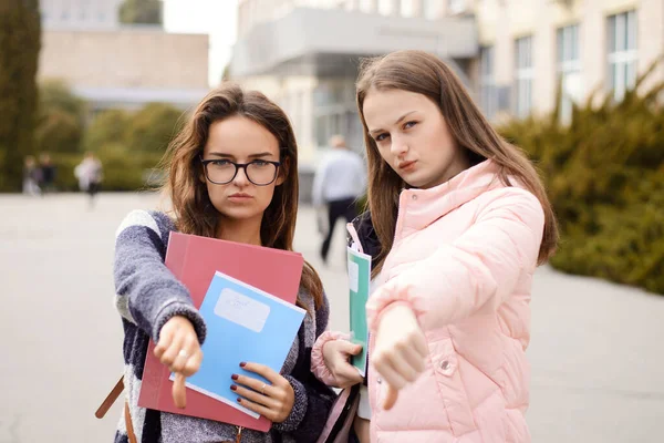Unhappy and regretful female students showing thumbs down in front of university building. Unhappy to study in university because of bad teaching stuff, lack of modern technologies and bullying