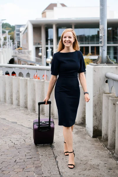 Smiling girl in black smart clothes at the railway station carrying big suitcase to the train