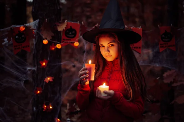 Spooky photo of small schoolgirl with red and white candle late in autumn forest. Halloween celebration, concept of witchcraft in forest