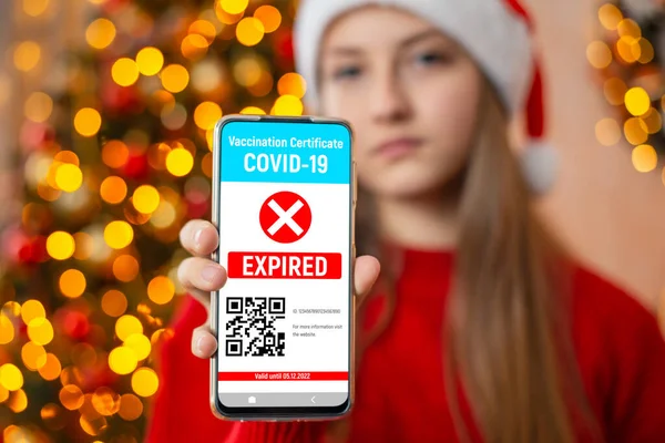 Smartphone with news about expired vaccination certificate on the screen, close up. Upset girl in Christmas clothes and garland lights on the background