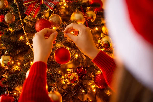 Back view of girls hands, hanging a red ball on the Christmas tree. Warm golden light, magical festive atmosphere