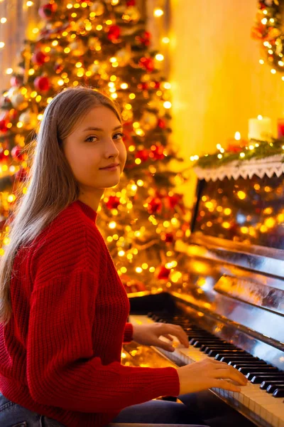 Horizontal shot of young female pianist playing her instrument near the decorated Christmas tree. Warm festive magical interior of the room, happy holidays concept