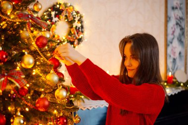 Young woman in red sweater decorates Christmas tree before winter holidays. Red and golden colours, warm light, new year and christmas concept