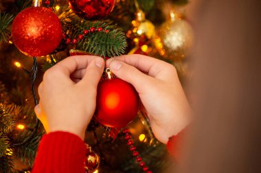 Winter holidays and decoration concept. Hands of a girl hanging red ball on Christmas tree, warm colours and mood