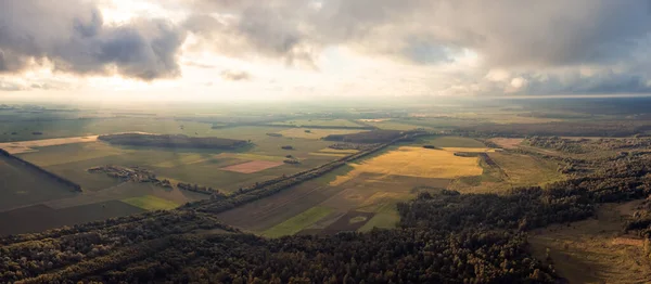 Wide panorama of flying high over countryside fields and forests. Dramatic sky during sunset, overcast sky with some places where sun rays come through thick clouds, drone point of view