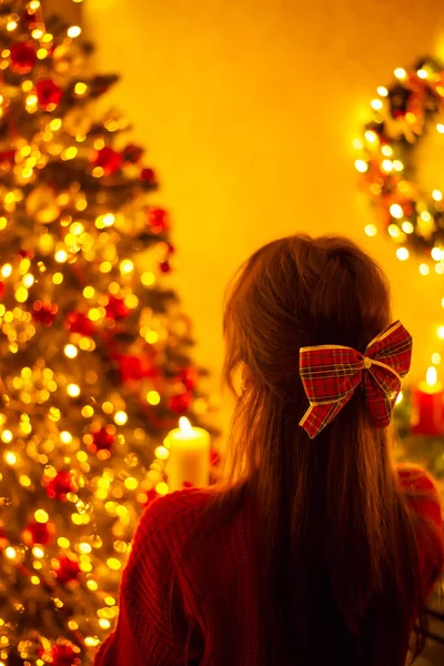 Christmas festive decorations, girl in decorated room for celebrating New Year with burning candle. Back shot of a dreamy beautiful girl admiring beauty of christmas lights in decorated room at home
