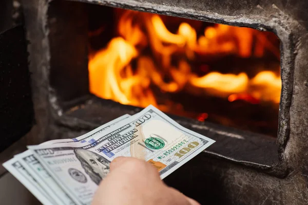 American money in hand of a person near opened door of solid fuel boiler, close up. Expensive heating of the house, warmth and comfort at home due to large expenses