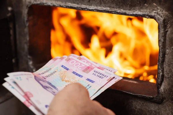 Burning money in solid fuel boiler as a symbol of high expenses for heating the house. Ukrainian hryvnia bills in hand of a man in front of bright flames inside the boiler