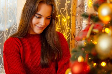 Portrait of a young cheerful girl in bright red sweater decorating Christmas tree. Christmas decorations and preparations concept