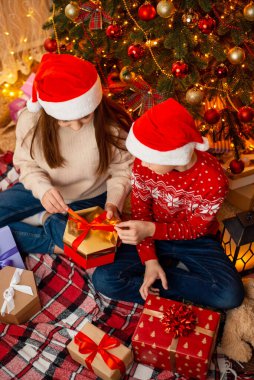 Horizontal photo of two children near the christmas tree, opening presents. Winter holidays, happy family atmosphere, festive interior.