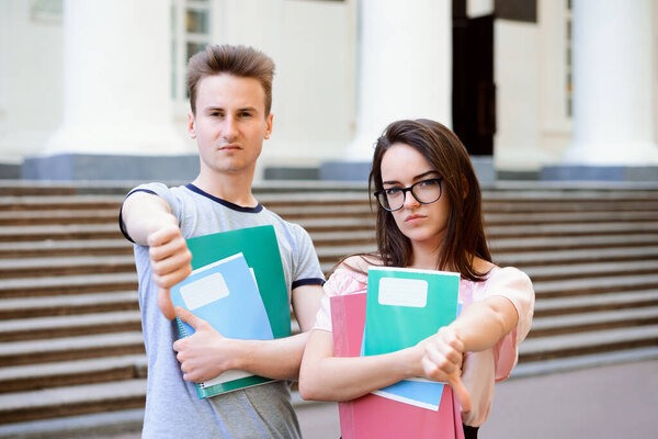 Грустное недовольство недовольных молодых студентов, показывающих большие пальцы, стоящих перед старым традиционным университетом. Концепция плохого обучения, плохой университет