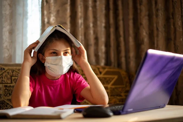 School girl in medical mask with a sad face at a desk working at laptop, sick and tired of constant learning, studying, doing tasks
