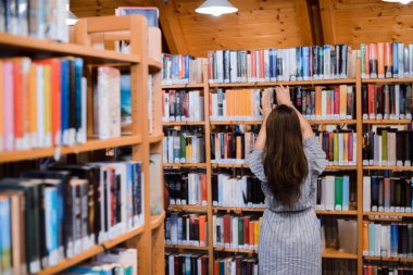 Back view of a girl with long beautiful hair standing near bookshelves looking for necessary book for her project in library
