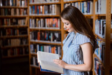 Picture of beautiful young brunette girl in bookstore standing near bookshelves, reading something in a book. Studying, learning with fun, buying books concept.