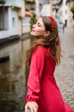 Pretty young brunette woman in bright rose dress and with wreath on her head standing on bridge in the romantic town; enjoys the moment, deep in her thoughts.