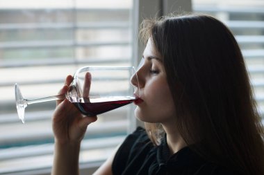 Beautiful brunette girl drinking bitter red wine. Lonely girl drinking wine at home, dreaming about something