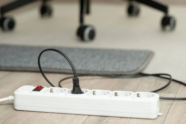 One black electric plug connects into white extension socket. Grey small carpet and wheels of an office arm-chair on the background