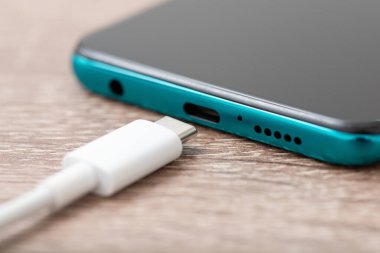 Smartphone of emerald colour and white usb Type C cable on the table, close up. Concept of charging the battery, up-to-date technologies for communication