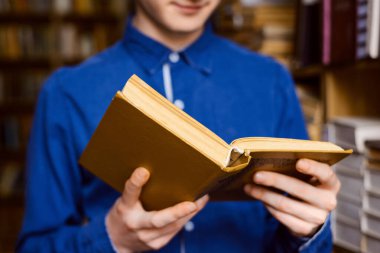 Close up picture of male student with a book in his hands, standing between shelves of books in the library. Young boy in blue shirt reading a book.