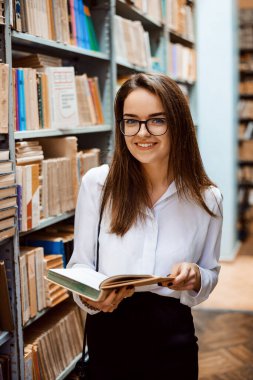 Beautiful young science worker in the library, holding an open book and looking to the camera smiling. Brunette girl in glasses, in white blouse and black skirt.