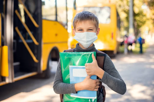 School learner in medical mask, holding learning materials standing near the school bus and showing thumbs up