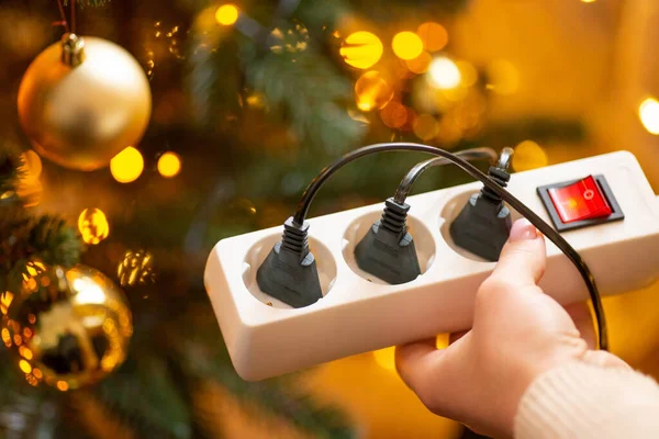 Close up of white multiple electrical socket in woman hands with three plugs in it, close up. Christmas tree with bright garland lights and golden baubles, home cozy atmosphere