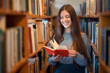 Ders arasında üniversitenin kütüphanesinde kitaplıkların arasında duran neşeli bayan öğrenci. Mutlu genç kadın okumayı, eğitim kavramını ve bilgi edinmeyi seviyor.
