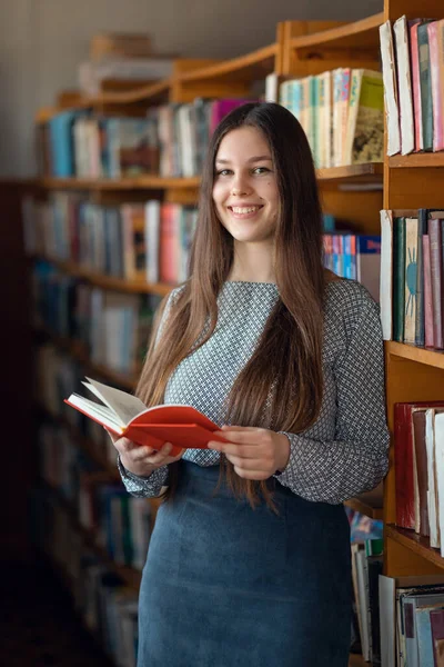本棚の近くに本を手に笑顔の少女 カメラを見てる 図書館 幸せな学生生活の概念 — ストック写真