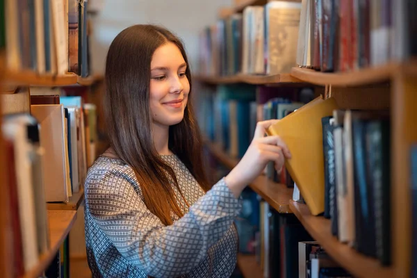 Молодая Улыбающаяся Девушка Стоящая Рядом Книжными Полками Библиотеке Берёт Книги — стоковое фото