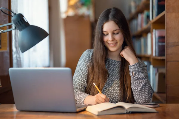 大学図書館で勉強している若い女性の笑顔 机に座ってノートを書き授業の準備ノートパソコンを使って — ストック写真