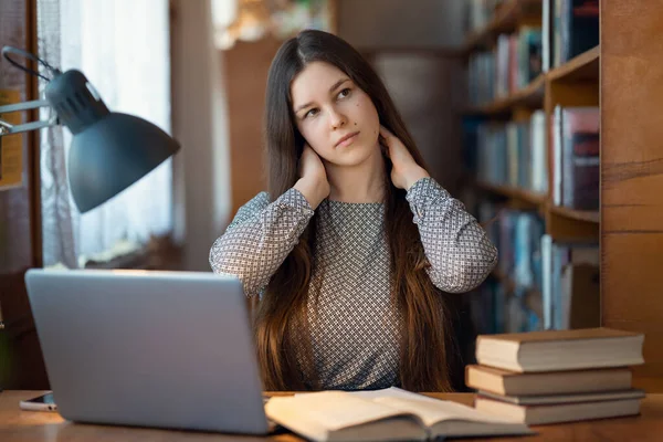 Young woman student is sitting at a table in the library, feeling tired, massaging her neck. Girl loaded with tasks, too much brainwork and not moving, neck pain