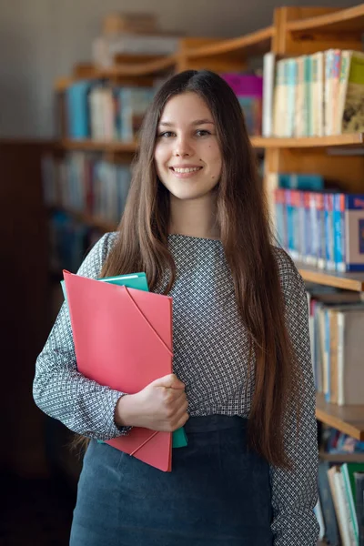 Young student girl in the library standing next to bookshelves, ready for learning. Education, knowledge and literature research concept, vertical shot, portrait