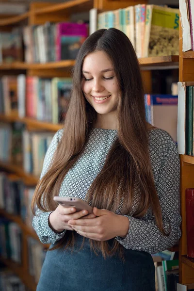 図書館にいる間に電話で笑顔の若い女の子は 友人と通信します 学生生活 大学図書館で勉強する女の子 携帯電話を使用して — ストック写真