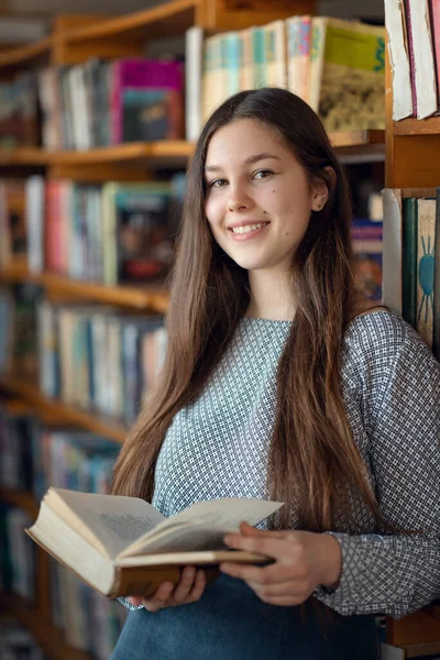 若い笑顔の女の子を見て読む 図書館の棚の近くに開いている本を持つ学生 読書のための文学を選択します — ストック写真