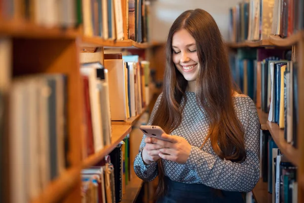 大学図書館にいる間 幸せな若い学生の女の子は友人からメッセージを受けました 携帯電話を使った女子学生の肖像画 — ストック写真