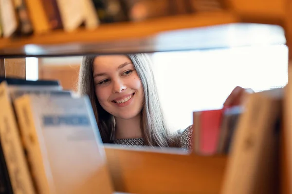 図書館の本の中で幸せな陽気な学生の女の子 文学を探している大学図書館の棚の間の少女の肖像画 — ストック写真
