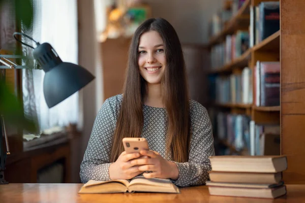 教室の机で携帯電話を使って働く若い白人の少女の笑顔 図書館で携帯電話の読書本を持つ幸せな女性学生 カメラを探して — ストック写真