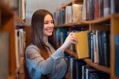 Kütüphanedeki kitaplıkların arasında kitap arayan gülümseyen esmer bir kız fotoğrafı. Bilgi edinmek, öğrenci hayatı, eğitim kavramları
