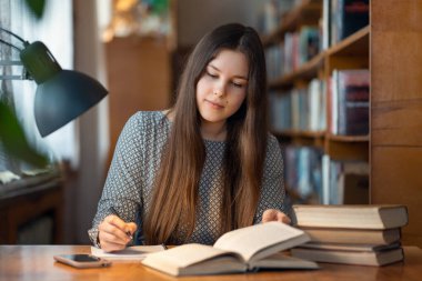 Konsantre olmuş genç kız öğrenci masada oturuyor, onun için bir şeyler okuyor ve yazıyor. Genç esmer kız kütüphanede çalışıyor, ödevlerini yapıyor.