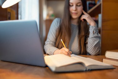 Çekici kız öğrenci ödevini bir masada yapıyor, işine odaklanmış. Kalem, dizüstü bilgisayar ve arka plandaki kitaplarla eline odaklanmış.