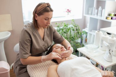 Profesyonel kozmetik uzmanı, güzellik salonundaki genç bayan müşterinin yüzüne soğuk hidrojenleşme için jel sürüyor. Derinin ultrasonik ve mekanik temizlik prosedüründen önce hazırlanması