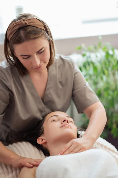 Face massage procedure at beauty spa salon, vertical shot, close up. Young woman getting massage treatment, female cosmetologist proceeding spa skin and facial beauty treatment