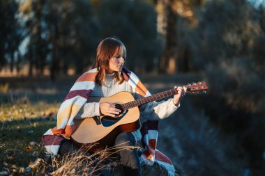 Genç bir kızın nehir kıyısında oturup gitar çaldığı atmosferik sonbahar akşamı çekimleri. Güz parkında gitar çalan güzel kız.