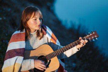 Nehir kenarında gitar çalan genç bir kızın portresi. Nehir kıyısında oturan ve gitar çalma, ustalaşma, yeni kompozisyon öğrenme eğitimi alan bir kız.